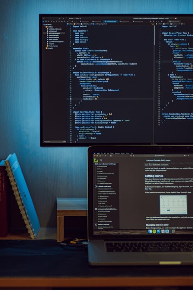 A desk at night with code open on a monitor and documentation on a laptop screen