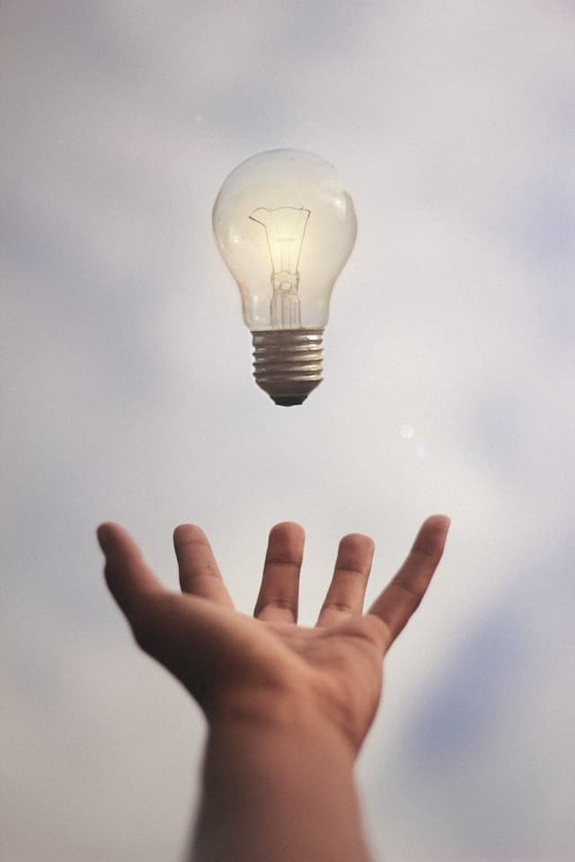 An open hand reaching up towards a lit light bulb floating against a pale sky